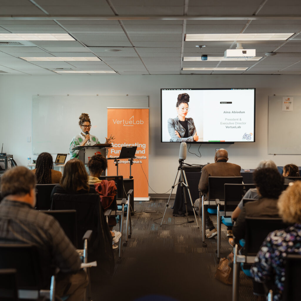 VertueLab President and Executive Director Aina Abiodun stands at a podium in front of an audience. There is VertueLab signage and a slide presentation next to her.